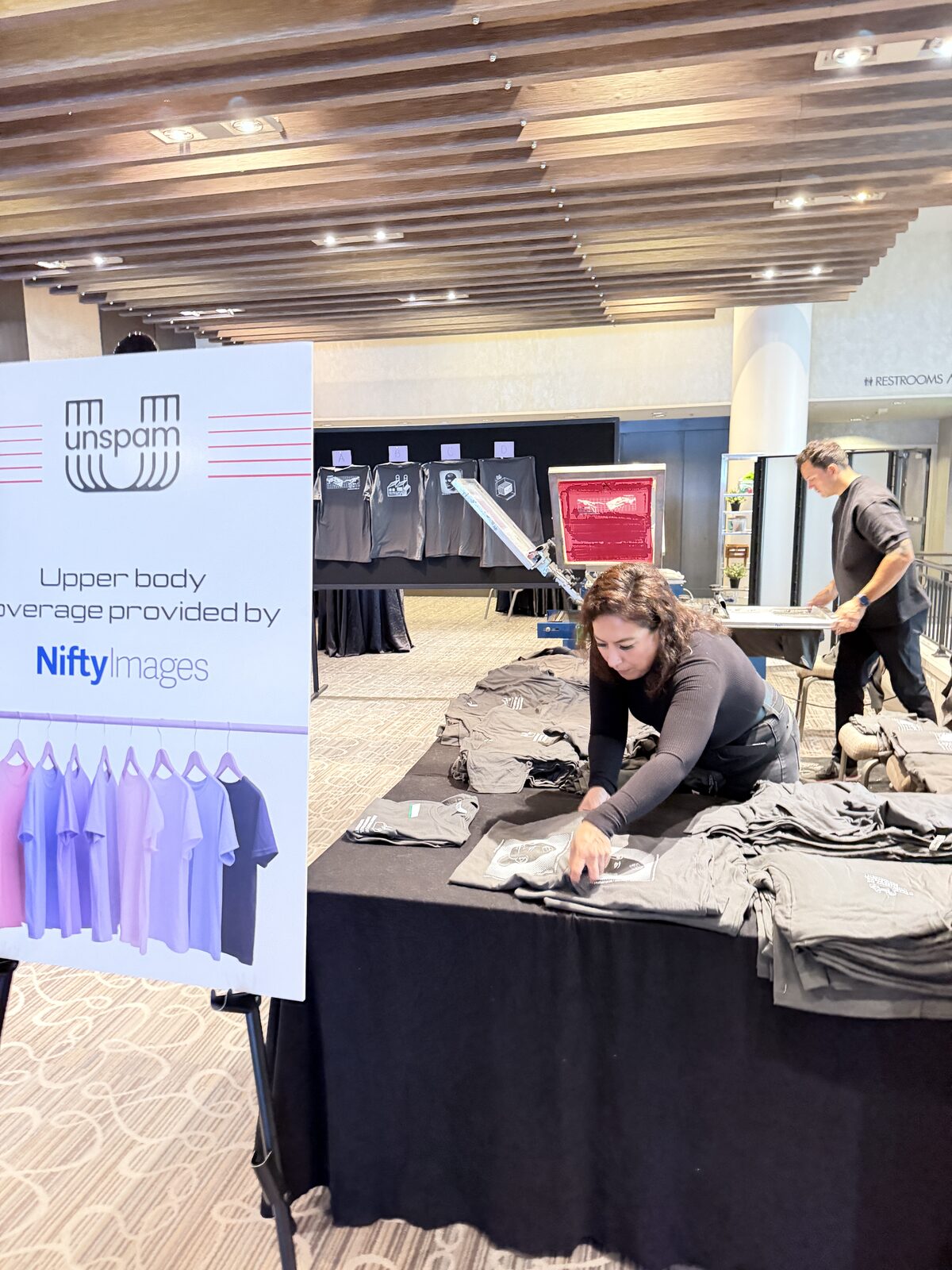 Folded shirts arranged in rows on a black merch table beside a conference welcome sign