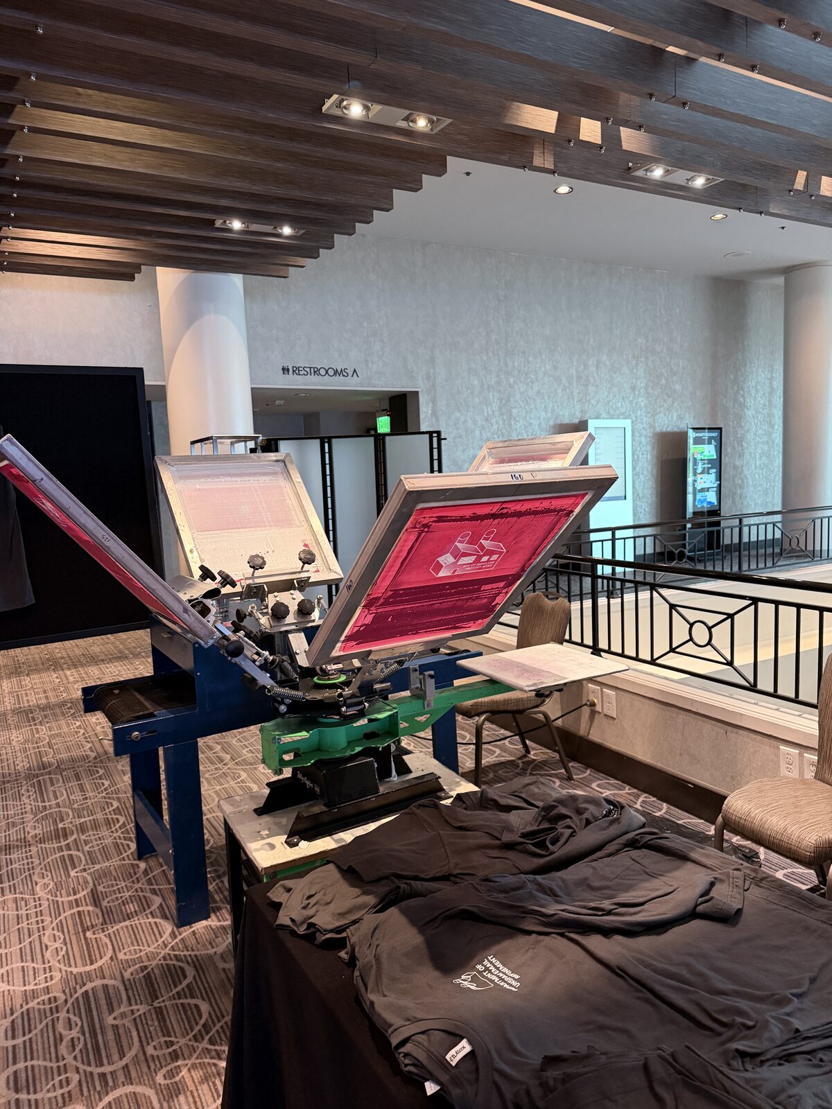 Four-color screen-printing press with red-tinted screens and stacked black shirts on a hotel mezzanine