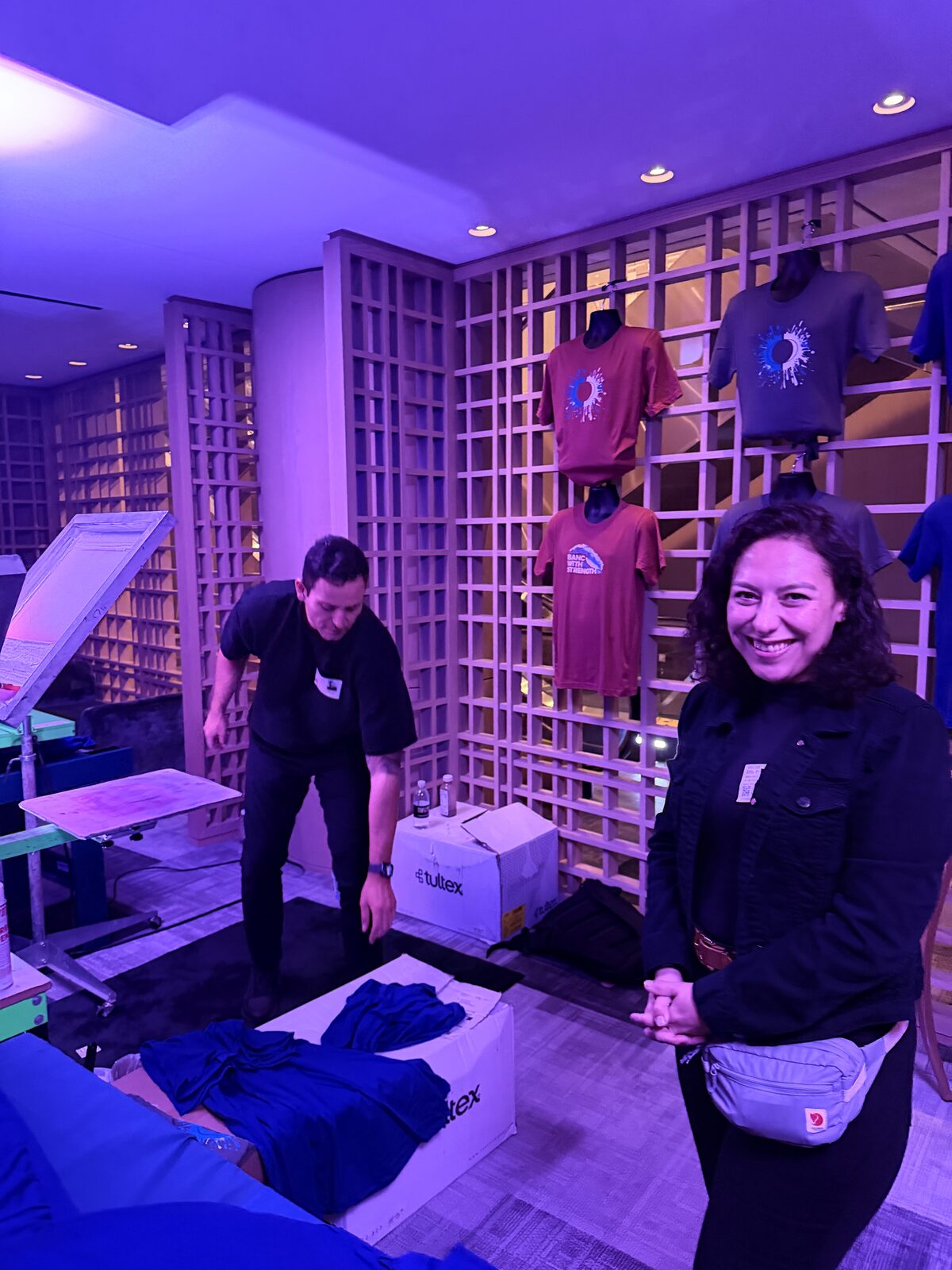 Print crew member at a purple-lit station with a shirt display wall, garment boxes, and a press behind
