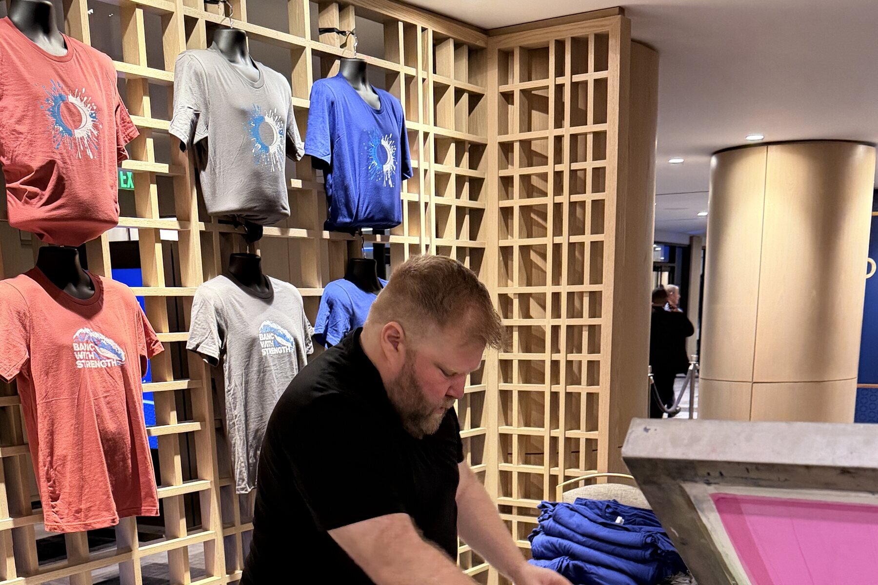 Printed tees on display busts in a wood lattice wall while a printer works at a press below